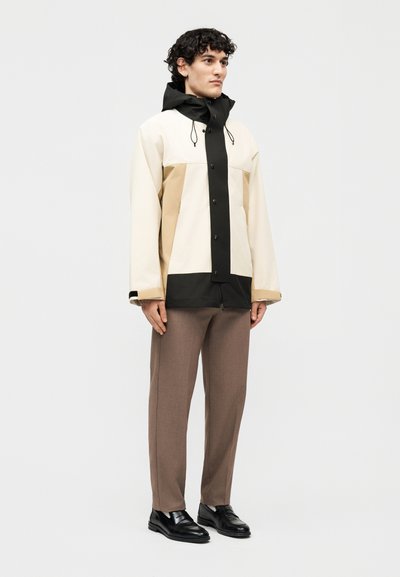 Άνδρας που φοράει χρωματιστό μπουφάν με κουκούλα σε μπεζ, κρεμ και μαύρο, καφέ παντελόνι και μαύρα παπούτσια, στέκεται μπροστά σε απλό φόντο.