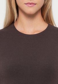 Ciemnobrązowa bluzka z okrągłym dekoltem, wykonana z gładkiego, teksturowanego materiału. Prosty design, dopasowany krój i brak widocznych wzorów czy akcentów.