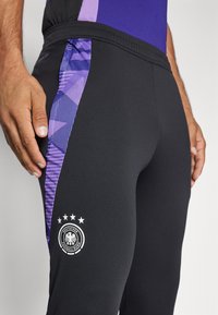 Pantaloni sportivi neri con pannello laterale viola texturizzato e logo con un'aquila tedesca con stelle sopra. Materiale morbido ed elastico.