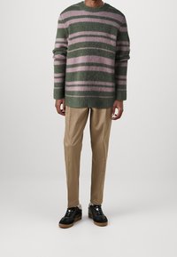 Pull rayé vert et rose en matière douce, associé à un pantalon beige et des baskets noires avec des semelles en caoutchouc. Manches longues et coupe décontractée.