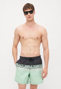 Shorts de bain noirs et vert menthe avec un cordon de serrage, présentant un grand logo imprimé sur le devant, fabriqués en tissu à séchage rapide.