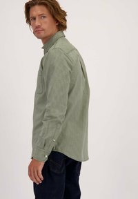 Man met schouderlang haar die omkijkt, gekleed in een lichtgroen gestreept overhemd met lange mouwen en donkerblauwe spijkerbroek tegen een witte achtergrond.