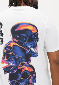 Mand iført en hvid t-shirt med tre farverige refleksive kranier trykt lodret på ryggen mod en ensfarvet baggrund.