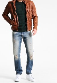 Giacca di pelle marrone, felpa verde con testo in rilievo, jeans blu strappati, sneaker bianche. Outfit casual con strappi e texture visibili.