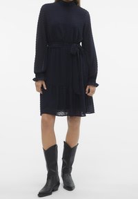 Marinblå klänning med texturerad tyg, långa ärmar, hög krage och knytning i midjan. Knälång med volangförsedda nederdelar. Bärs med svarta cowboystövlar.