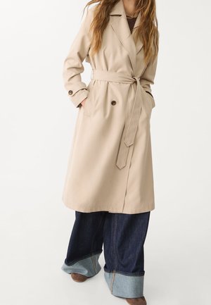 Beige trenchcoat met een tailleband, twee voorzakken, knoopaccenten en een kraag. Ggedragen over brede donkerdenim jeans.