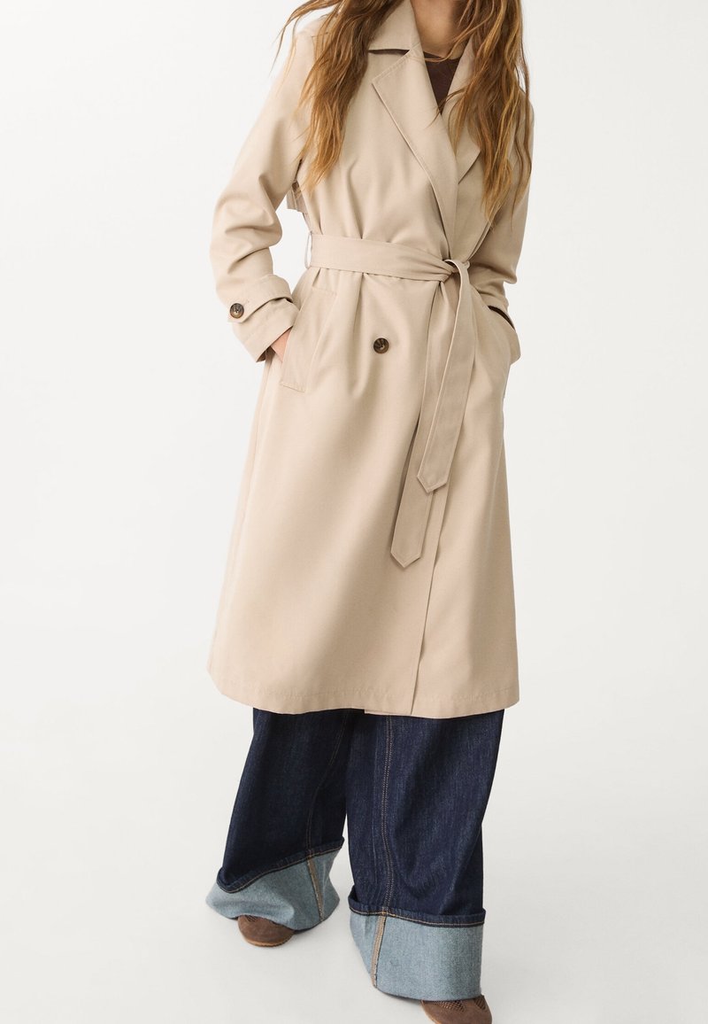 Beżowy płaszcz trencz z paskiem w talii, dwiema przednimi kieszeniami, guzikami oraz kołnierzykiem. Noszony na szerokie ciemne dżinsy.