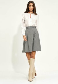 Kobieta w białej bluzce z długimi rękawami, czarno-białej spódnicy w wzór w zęby psa, oraz beżowych kozakach na wysokich obcasach do kolan, stojąca na tle jednolitego tła.