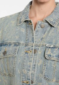 Jeansowa kurtka w jasnoniebieskim kolorze, z wyblakłym praniem, przetartą teksturą, srebrnymi detalami i tkaną metką na kieszeni piersiowej.