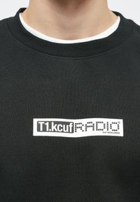 Μαύρο φούτερ με λαιμόκοψη, διακοσμημένο με μια λευκή ορθογώνια ετικέτα που φέρει το κείμενο "T1.kcuF RADIO" και "HUF WORLDWIDE" σε μαύρα γράμματα.