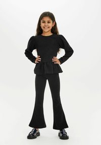 Črni dvodelni komplet, ki vključuje top s peplum krojem z dolgimi rokavi in razširjene legice, izdelan iz teksturirane tkanine, v kombinaciji s sijajnimi črnimi čevlji.