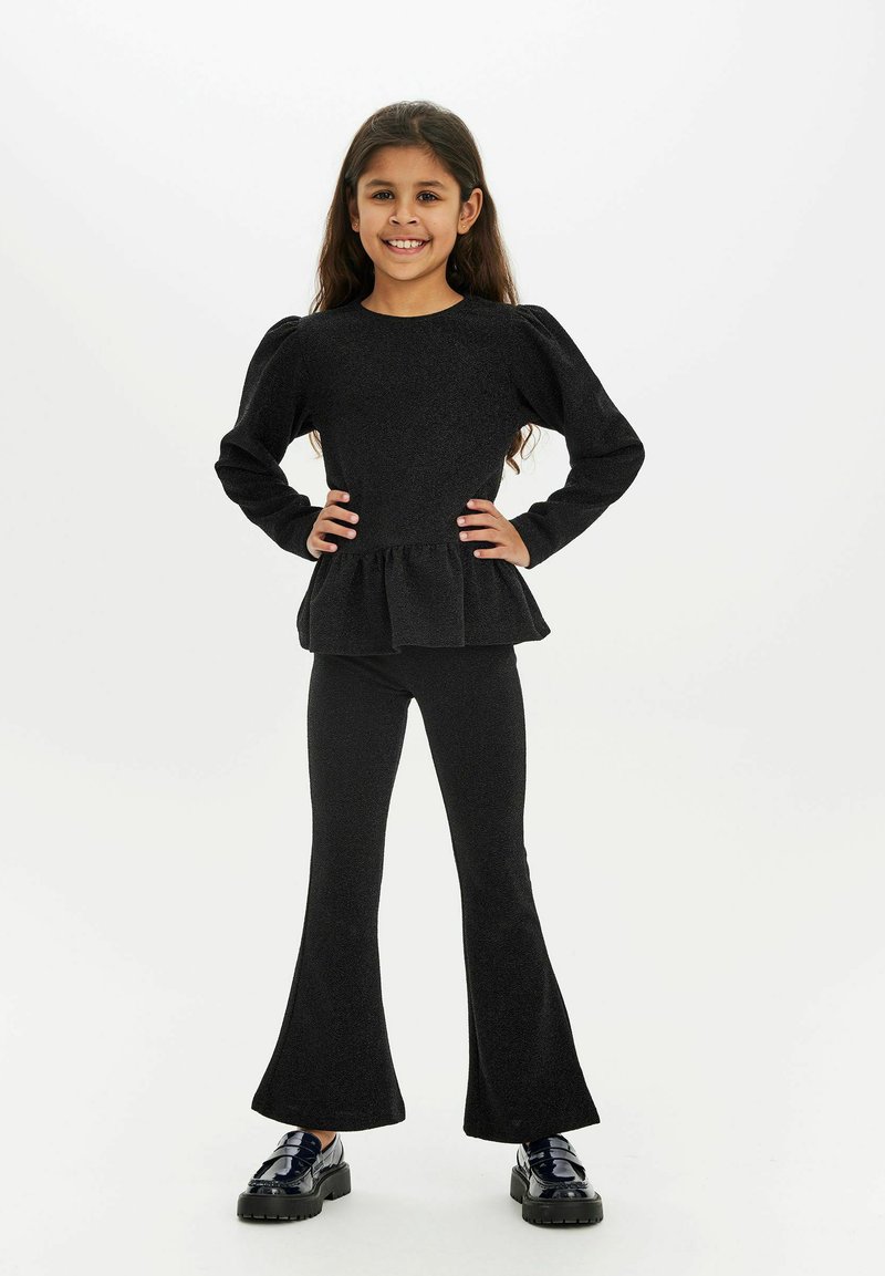 Črni dvodelni komplet, ki vključuje top s peplum krojem z dolgimi rokavi in razširjene legice, izdelan iz teksturirane tkanine, v kombinaciji s sijajnimi črnimi čevlji.