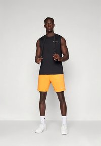 Camiseta deportiva sin mangas negra y pantalones cortos naranjas, ambos de tela ligera. Zapatillas blancas y una cuerda para saltar en la mano.