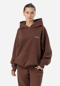 Felpa marrone con vestibilità rilassata, cappuccio con cordino, tasca frontale e discreto logo "Pеgador" sul petto. Tessuto morbido e strutturato.
