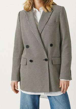 Femme portant une chemise blanche, un blazer croisé pied-de-poule noir et blanc, et un jean bleu sur fond uni.