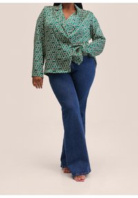 Grün gemusterte Bluse mit geometrischem Design, langen Ärmeln und einer Schleifendetail an der Taille, kombiniert mit blauen Schlagjeans.