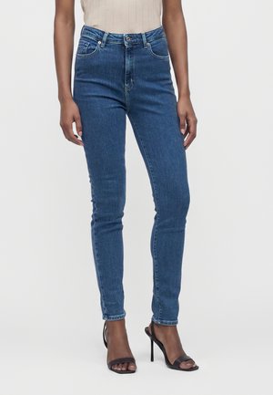 Μπλε τζιν skinny fit φορεμένα με μαύρα σανδάλια ψηλοτάκουνα και ένα μπεζ ριμπ ασύμμετρο αμάνικο τοπ, δείχνοντας από τη μέση μέχρι τα πόδια.