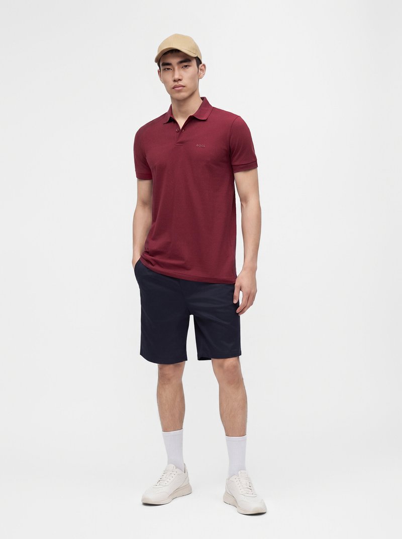 Jeune homme debout portant une casquette beige, un polo bordeaux, un short foncé, des chaussettes blanches et des baskets blanches sur un fond clair uni.