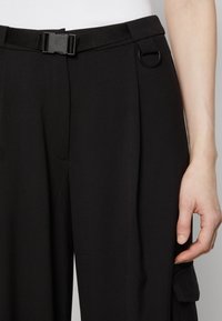 Black trousers feature a sleek design with a belt and loop detail, pleats, and a side pocket. Made from a smooth material, showcasing a minimalist style.