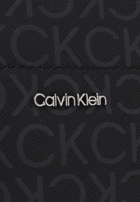Tissu noir texturé présentant un motif en relief subtil avec les lettres "CK". Logo "Calvin Klein" argenté affiché de manière proéminente.