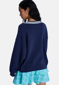 Μπλε ναυτικό, φαρδύ πουλόβερ με υφή και γιακά με λευκές ρίγες, συνδυασμένο με πολύστιχη φούστα σε ανοιχτό μπλε και πράσινο καρό.