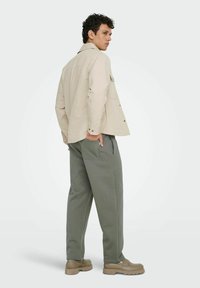 Homme portant une veste beige, un pantalon décontracté gris et des chaussures couleur sable, debout avec une main dans la poche arrière, regardant sur le côté sur un fond uni.