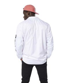 Camicia bianca a maniche lunghe con una vestibilità rilassata, caratterizzata da un design semplice e dettagli neri sulle maniche. Indossata con un cappellino rosa.