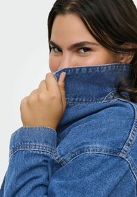 Džinsinis švarkas su mėlynu atspalviu, sulankstytu apykakle ir matoma siūle. Modelis laiko apykaklę ir dalinai sukasi, demonstruodamas tekstūrą ir tinkamumą.