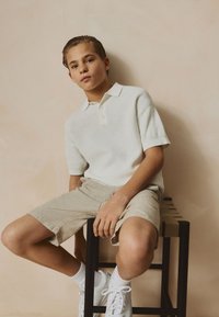 Short-sleeve white polo shirt with textured pattern, beige linen shorts, sitting on a black stool, wearing white sneakers with ankle socks.
