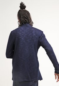 Námořnická modrá lehká košile s dlouhým rukávem a texturovanou tkaninou, s vysokým límcem, volným střihem a zakřiveným spodním lemem, viděná z zadní strany.