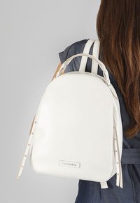 Zaino in finta pelle bianca con forma arrotondata, doppio manico e spallacci regolabili. Presenta un logo e accenti decorativi sui lati.