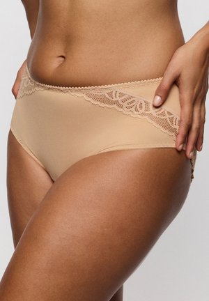 Vrouw met beige ondergoed met kanten rand, hand op de heup, waarbij het bovenlichaam en de bovenkant van het been zichtbaar zijn tegen een effen achtergrond.