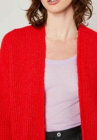 Rdeča pletena jopica z teksturiranim zaključkom, odprta spredaj in z dolgimi rokavi, nosi se čez svetlo vijolično prilegajočo majico. Vidna je črna pasnica.