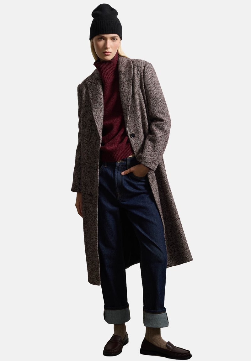 Lange, tweed jas in bruintinten, kastanjebruine gebreide trui, donkere denim jeans met opgerolde boorden, zwarte beanie en bruine loafers.