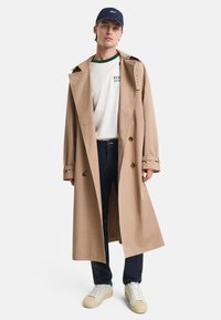 Beige trench coat with a double-breasted design, large lapels, and button accents, worn over a white shirt with green trim and dark pants.