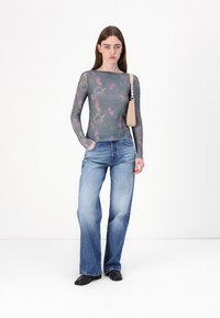 Long-sleeve mesh top in gray with colorful abstract patterns, paired with flared blue jeans and black slip-on shoes. Minimal beige shoulder bag.