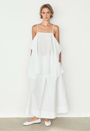 Femme aux cheveux courts portant une robe blanche ample sans manches à couches et des ballerines blanches, debout devant un fond uni.