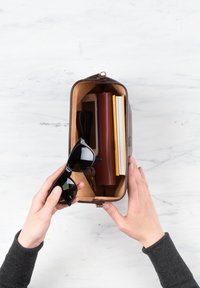 Mani che tengono una borsa marrone aperta contenente occhiali da sole neri, smartphone, chiavi, portafoglio marrone e due quaderni su una superficie bianca.