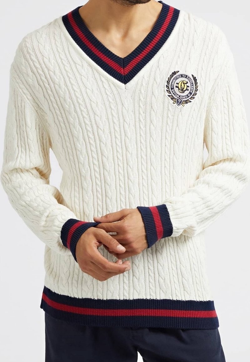 Homme portant un pull à col en V en maille torsadée blanche avec des rayures bleu marine et rouges sur le col, les poignets et l'ourlet, et un blason brodé sur la poitrine.
