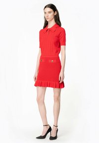 Abito rosso lavorato a maglia con colletto polo a maniche corte, bottoni decorativi e orlo arricciato; abbinato a scarpe con tacco alto nere.