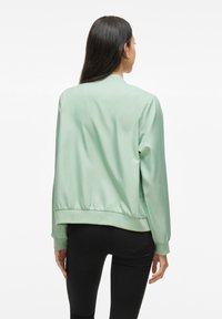 Giacca bomber in satin verde chiaro con cintura e polsini aderenti. Presenta un colletto semplice e una superficie liscia, mostrata da dietro.