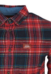 Camisa de franela a cuadros en rojo y azul marino con cuello botón, que incluye botones marrones y un parche con logo en el pecho, fabricada con tejido texturizado.