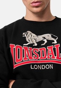 Felpa in cotone nera con un grande leone ricamato e la scritta "LONSDALE LONDON" in rosso e bianco sul davanti. Design classico con collo a giro.