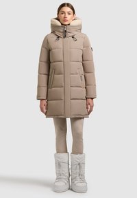 Frau steht und trägt einen beigefarbenen, gepolsterten Wintermantel mit hohem Kragen und weißen Schneestiefeln vor einem einfarbigen Hintergrund.