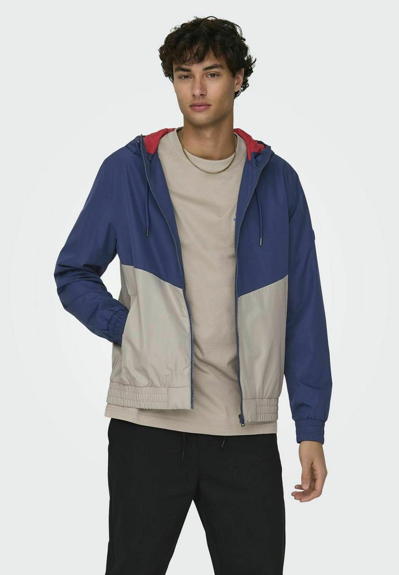 Kleurrijke windbreaker in marineblauw en lichtgrijs met een rode capuchon. Bevat elastische boorden, ritsluiting en een gevoerde capuchon. Lichtgewicht stof.
