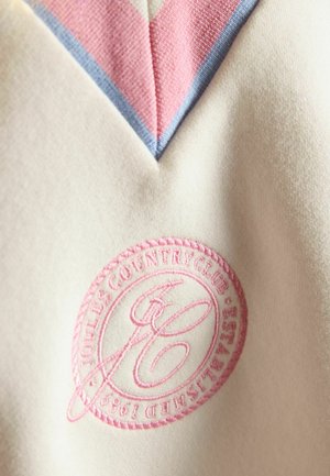 Kermanvärinen vaatekappale, jossa on vaaleanpunainen ja sininen V-päänti koristeluna sekä vaaleanpunainen kirjailtu pyöreä logo, jossa lukee "Joules Country Club Established 1989".