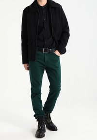 Veste noire zippée avec un col en shearling, chemise noire à boutons, pantalon vert et bottes de combat noires. Tissu texturé et coupe ajustée.