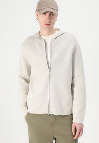 Ljusgrå zip-up hoodie i mjukt tyg som har en huva, framdragkedja och långa ärmar. Matcher med olivgröna byxor.