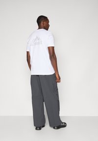 T-shirt bianco a maniche corte con grafica di montagna e logo "TERREX" sul retro; abbinato a pantaloni cargo grigi scuri e scarpe nere.