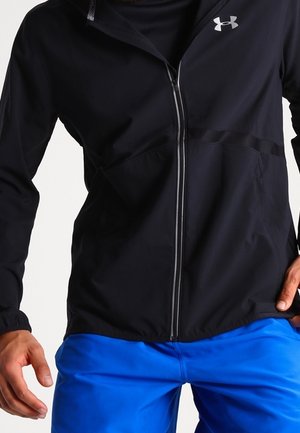 Czarna kurtka na zamek błyskawiczny z logo Under Armour na piersi, noszona z jasnoniebieskimi spodniami, ręce swobodnie opuszczone wzdłuż ciała na białym tle.